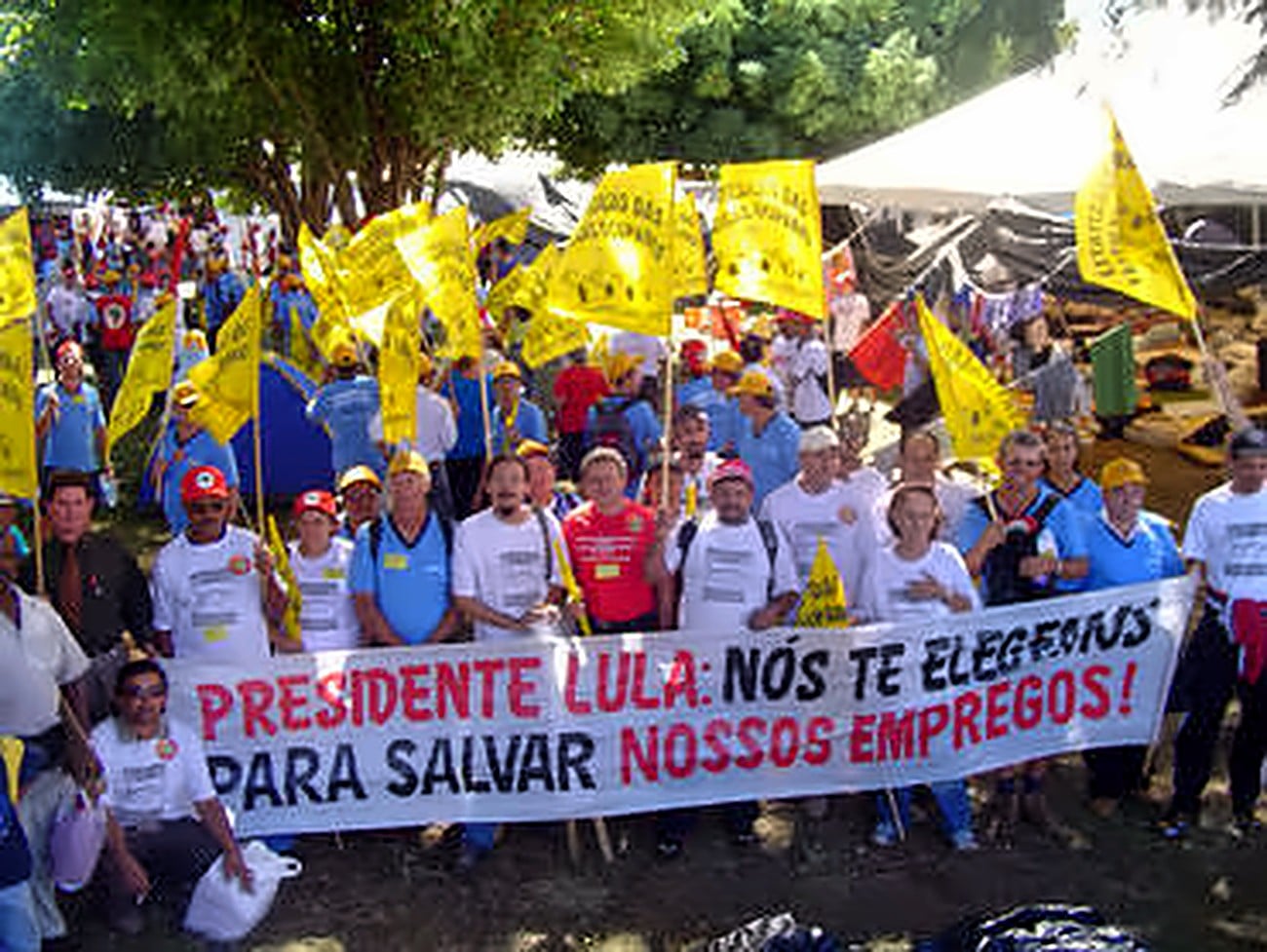 Trabalhadores com bonés e bandeiras amarelas atrás da faixa Presidente Lula: nós te elegemos para salvar nossos empregos!