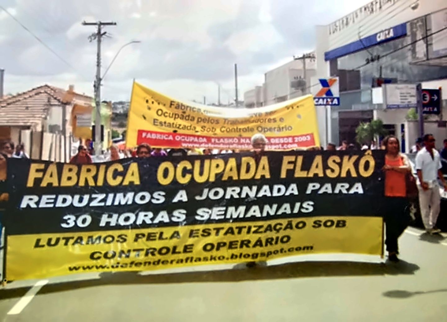 Passeata na rua com a faixa preta e amarela Fábrica Ocupada Flaskô: reduzimos a jornada para 30 horas semanais, lutamos pela estatização sob controle operário. Atrás, outra faixa amarela da fábrica ocupada.